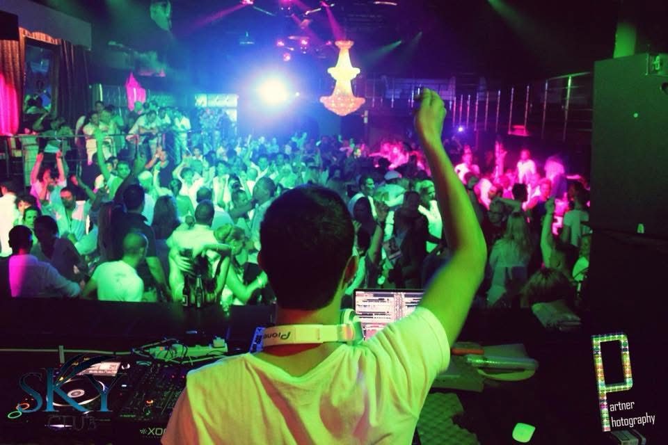 Packed club dancefloor in neon wash, seen from behind the decks